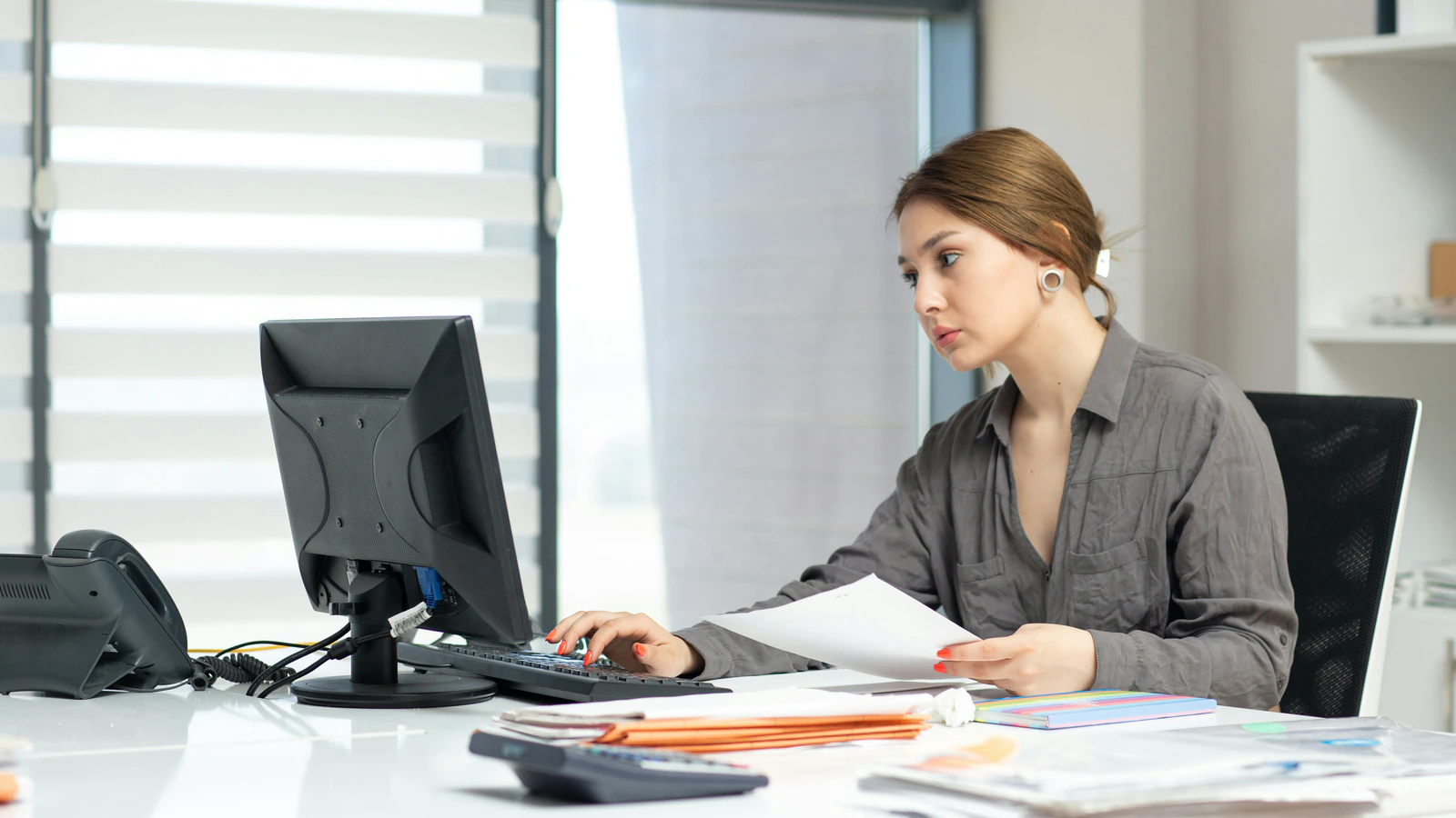An office worker at a desk full of files, paperwork, a phone and a calculator, working through admin by hand.